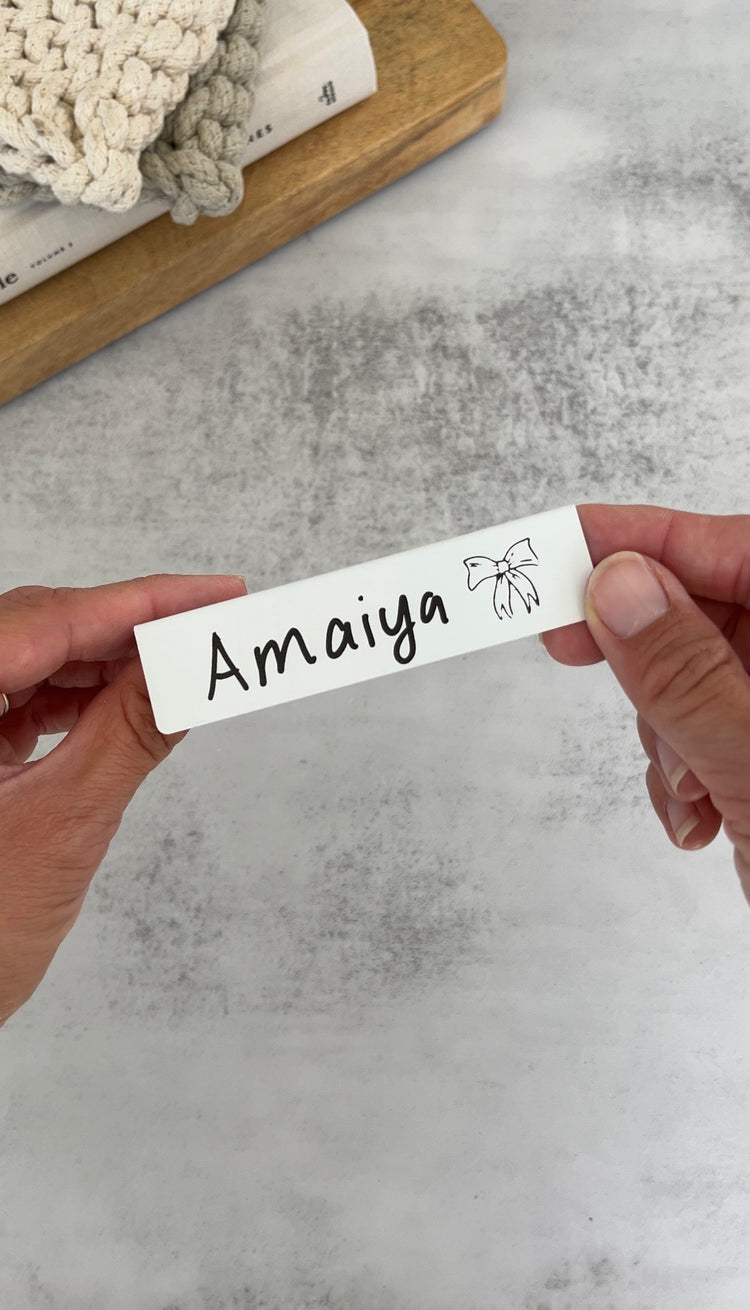 Hands holding a white TidyPop place card that says Amaiya with a bow