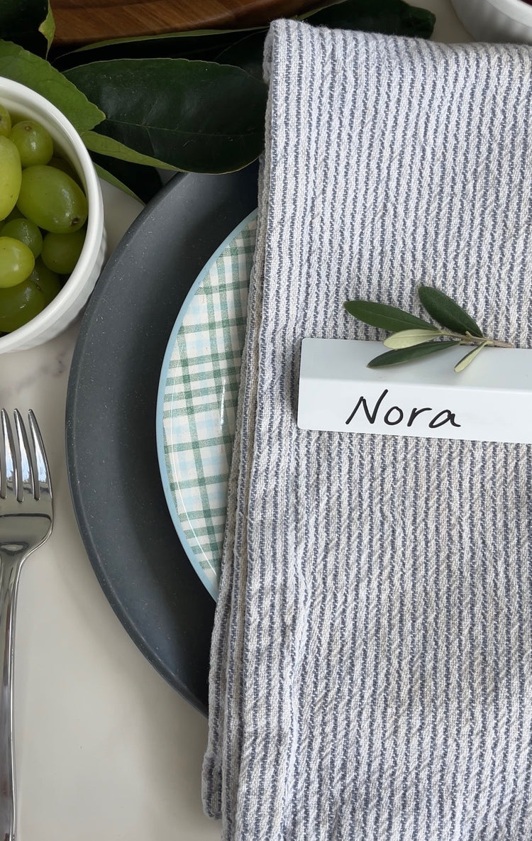 A white TidyPop place card reading Nora on a striped linen napkin