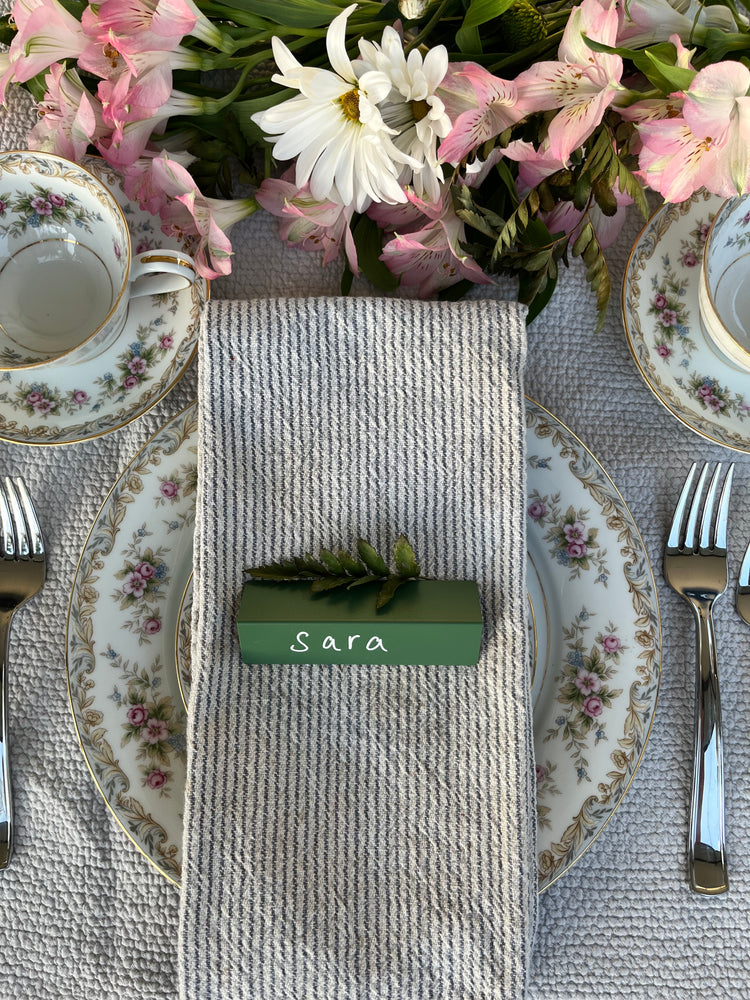 A sage TidyPop place card reading Sara on a linen napkin with floral china
