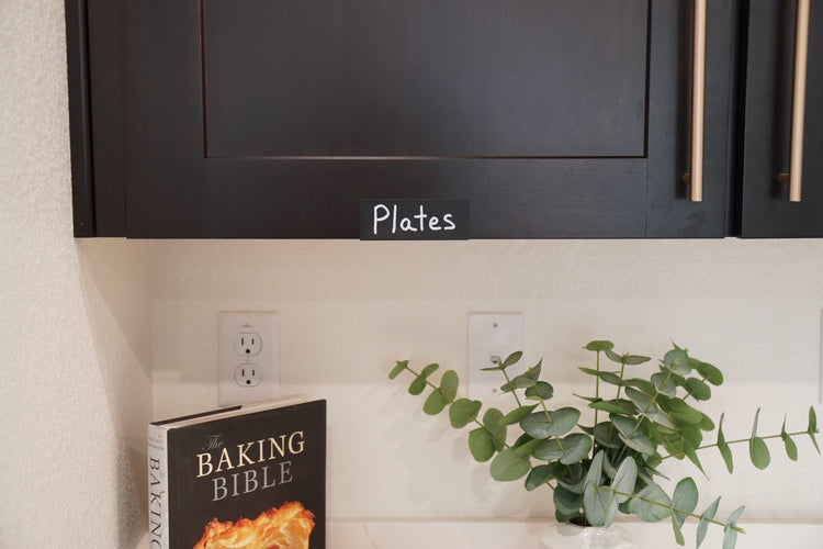 Black TidyPop clip reading Plates on a black kitchen cabinet above a cookbook and eucalyptus